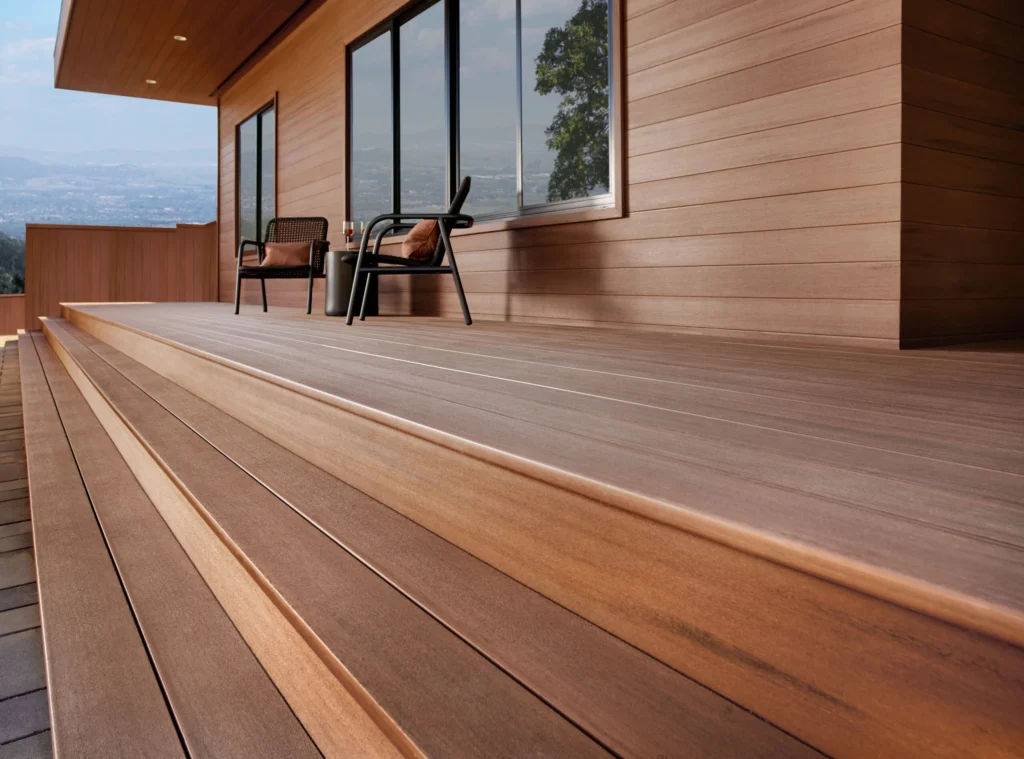 A modern home with TimberTech Mahogany porch boards, featuring wide steps and sleek horizontal paneling. The rich reddish-brown tone of the boards creates a smooth transition from the steps to the main deck, complementing the surrounding wood-clad walls and black-framed glass windows.