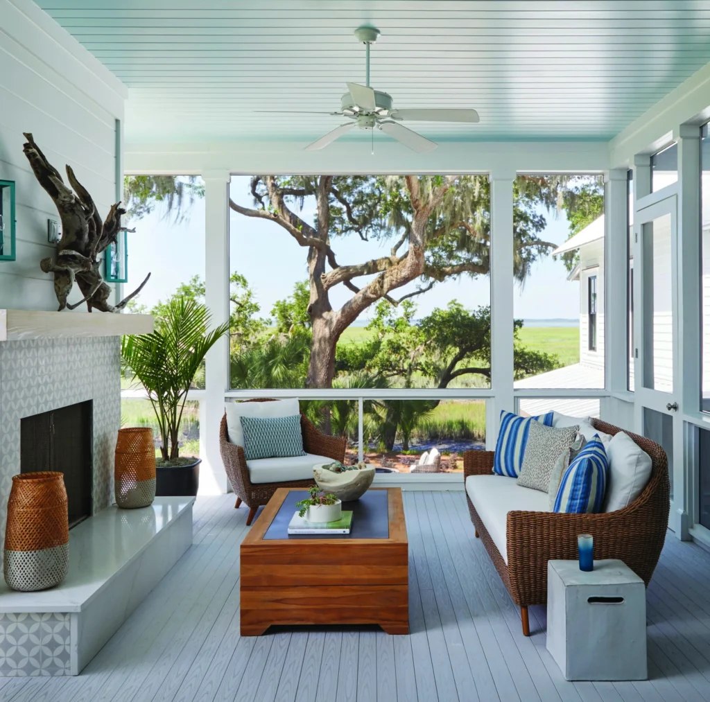 A bright, coastal-style screened porch showcases TimberTech Porch in Oyster, creating a breezy and inviting retreat. The cool-toned boards feature realistic woodgrain texture and a smooth, matte finish that complements the wicker furniture and blue accents. Large windows frame the surrounding trees and waterfront, letting natural light bounce off the pale decking surface.