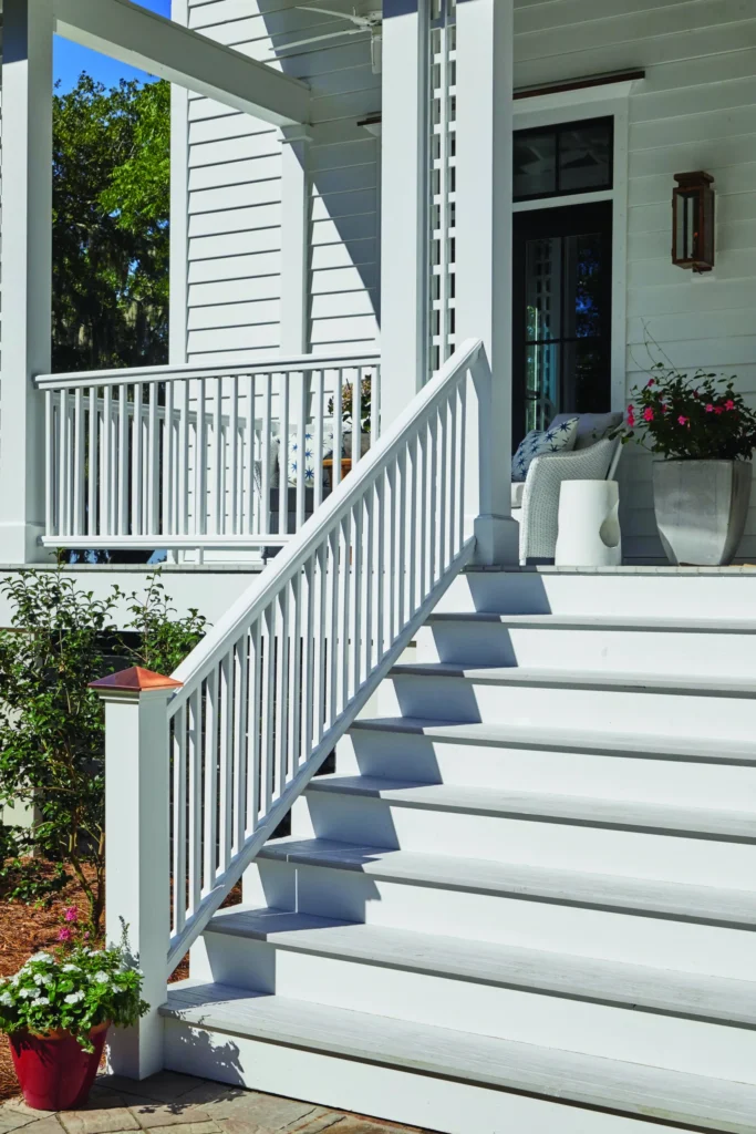 A front porch stairway features TimberTech Porch in Oyster decking paired with crisp white railing and trim. The subtle gray color and refined woodgrain pattern create a seamless extension of the home's coastal exterior. Pops of greenery and copper accents on the post caps complete the welcoming entryway.