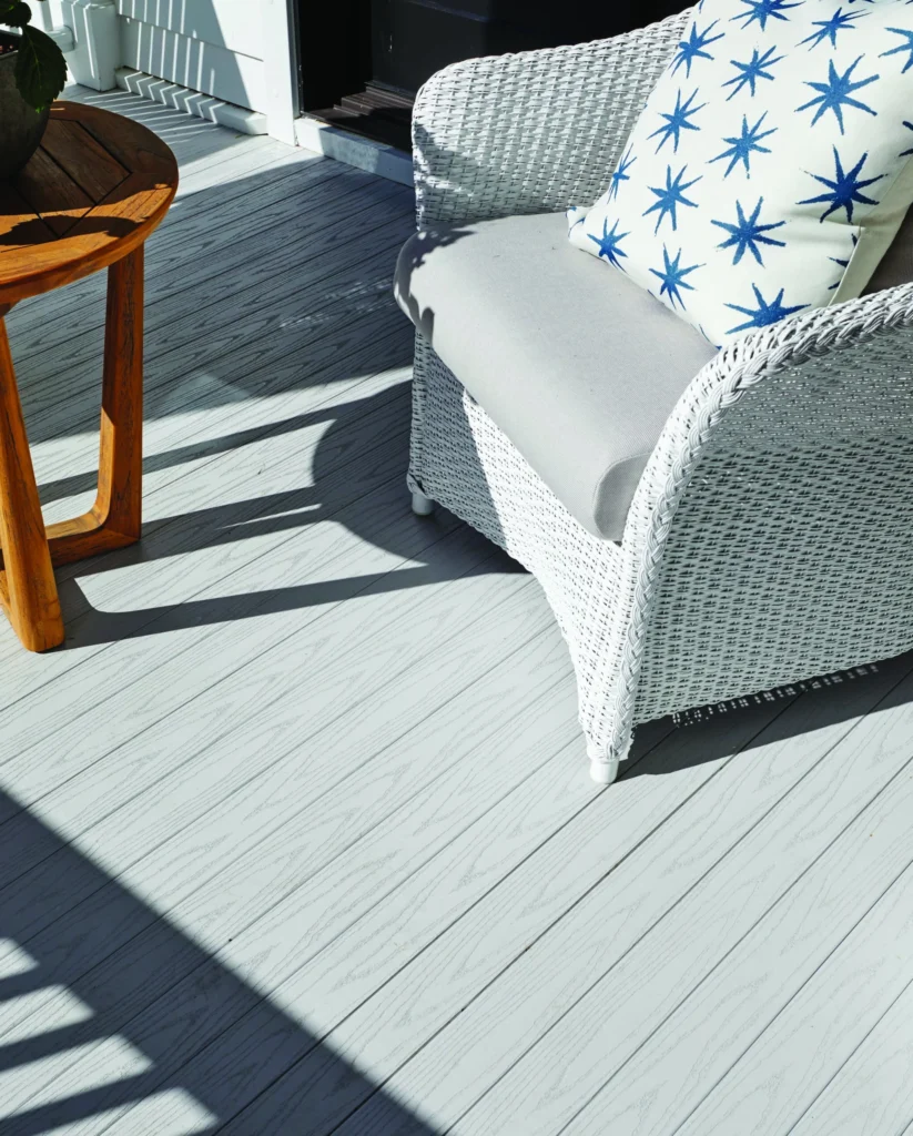 Close-up of TimberTech Porch in Oyster decking beneath a white wicker chair with a star-patterned cushion. The soft gray boards offer a natural woodgrain appearance and a cool undertone that contrasts beautifully with the cozy, coastal seating. Sunlight highlights the clean lines and texture of the tongue-and-groove design.