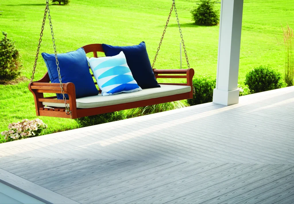 A wooden porch swing with blue and striped cushions hangs above a TimberTech Porch deck in Oyster color. The tongue-and-groove boards feature a subtle woodgrain texture in light gray, arranged in a chevron pattern for a classic and airy coastal look. The lush green lawn in the background adds contrast to the soft, cool-toned decking.
