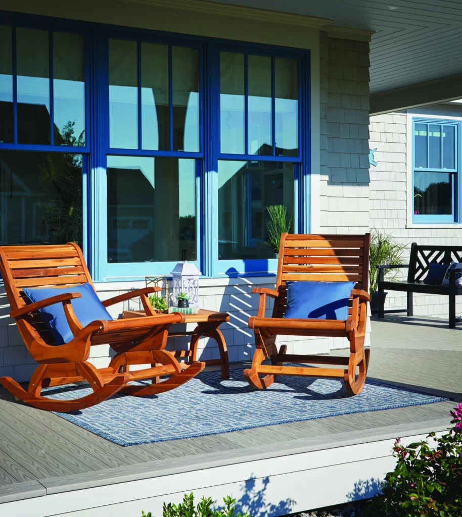 Two wooden rocking chairs with blue cushions sit atop a TimberTech Porch Slate Gray deck, paired with a small coffee table and decorative lantern. The tongue-and-groove decking boards showcase a dark gray hue with woodgrain detailing, framed by light trim for added contrast. Bright blue window frames in the background create a vibrant and beachy aesthetic.