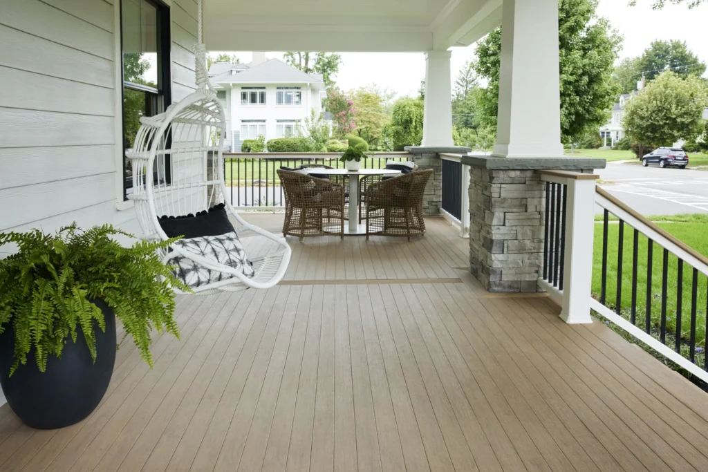 Spacious porch wrapped in warm Weathered Teak PVC decking with a white hanging chair, potted fern, and woven dining set under classic white pillars and stone accents.
