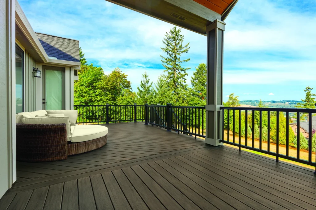 A spacious elevated deck made with TimberTech Prime+ Dark Cocoa composite decking, featuring rich brown boards with natural woodgrain textures. The deck overlooks a scenic landscape of trees and distant hills, framed by a modern black metal railing and shaded by a sleek patio roof. A round wicker daybed with cream cushions sits in the corner, creating a cozy outdoor retreat.