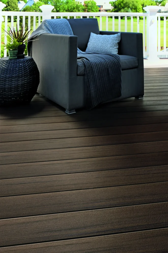 Close-up of TimberTech Prime+ Dark Cocoa decking on a sunlit porch, highlighting the realistic woodgrain detail and subtle color variation. A contemporary armchair with dark gray upholstery and a throw blanket rests beside a black wicker side table and a small potted plant, creating a peaceful reading nook.