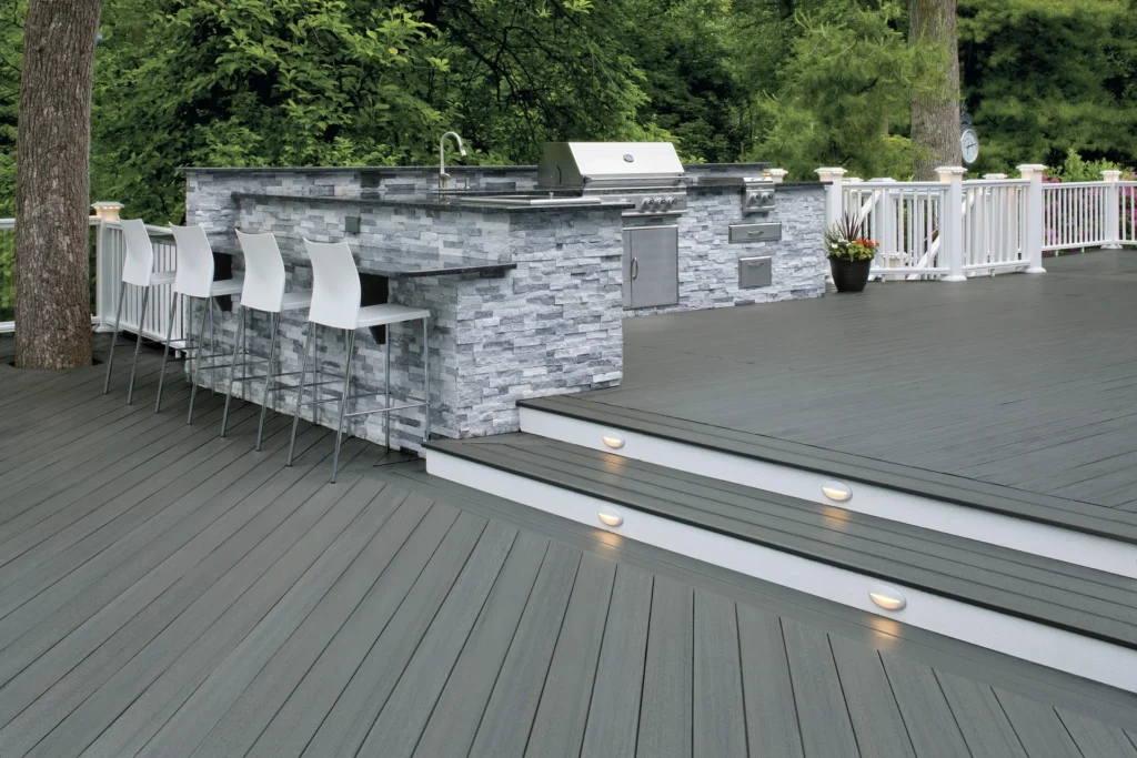 An elevated backyard deck built with TimberTech Prime+ Sea Salt Gray boards, featuring a modern outdoor kitchen clad in stacked gray stone and stainless steel appliances. The cool-toned decking complements the white railings and gray countertops, creating a cohesive, contemporary space.