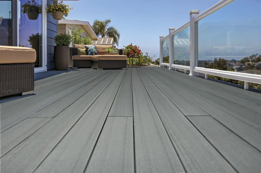 A spacious rooftop deck showcasing TimberTech Prime+ Sea Salt Gray decking paired with wicker patio furniture, potted plants, and glass railings. The soft, ocean-inspired gray tone blends beautifully with the blue house siding and bright coastal sky in the background.