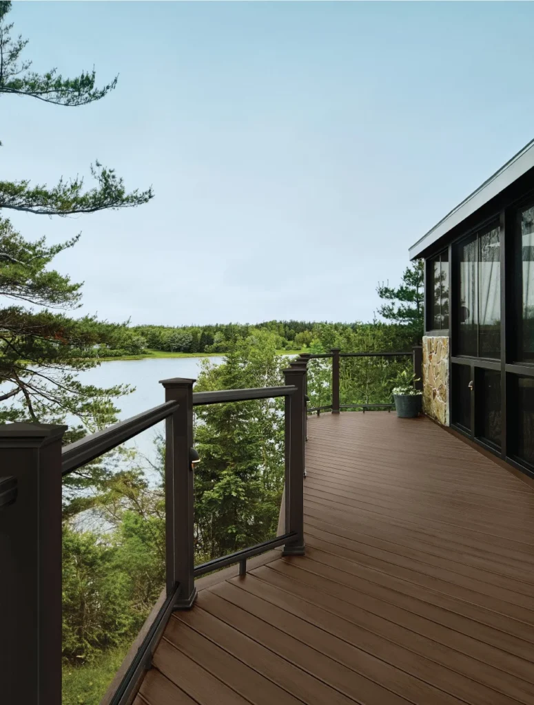 A scenic outdoor deck built with TimberTech Terrain+ Dark Oak composite decking boards, overlooking a tranquil lake surrounded by lush greenery. The deck features clean board lines and a matte woodgrain texture, framed by black aluminum and glass railing, creating a sleek yet natural look ideal for elevated or waterfront views.