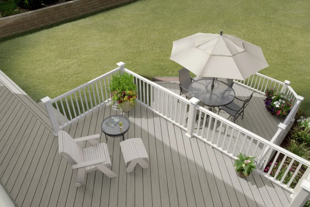 Overhead view of a TimberTech Terrain+ Weathered Oak deck featuring light gray composite decking with a natural wood grain finish, white railing, and outdoor furniture including a round table with an umbrella and Adirondack chairs - surrounded by a well-manicured green lawn and flower planters.