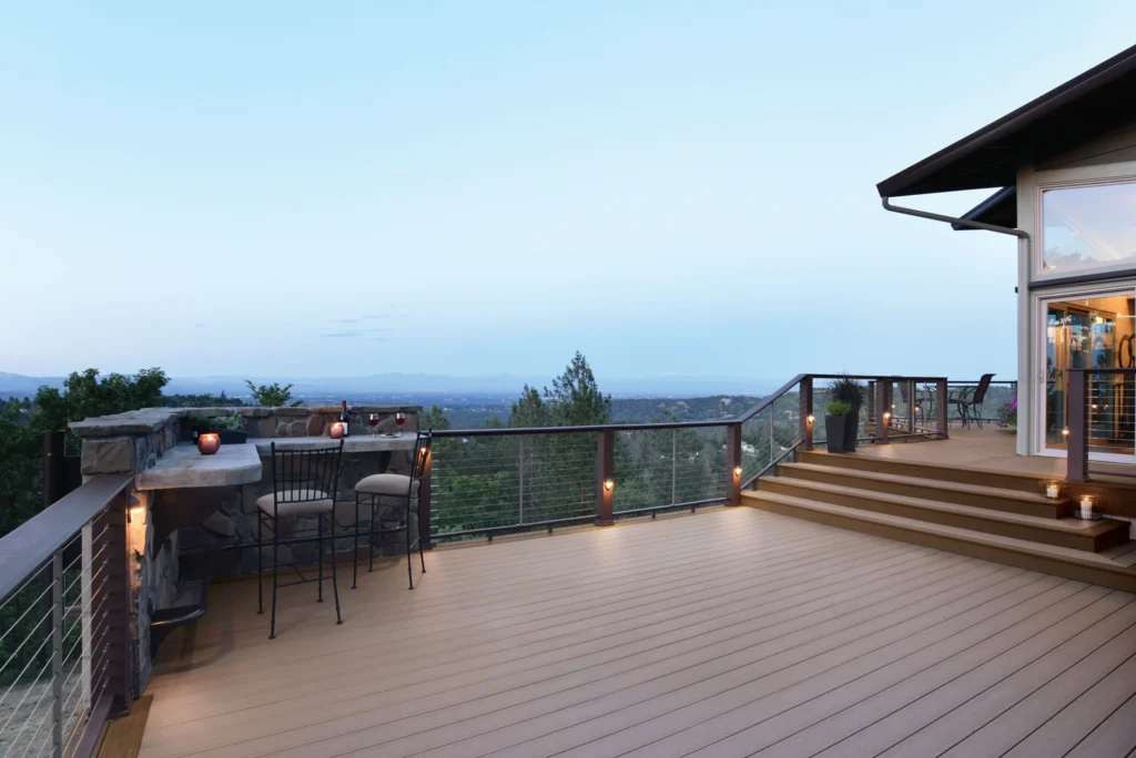 Spacious outdoor deck featuring TimberTech Terrain Sandy Birch decking, surrounded by modern cable railing and scenic mountain views at sunset; the light brown composite boards create a soft, inviting contrast with the dark railing and built-in stone bar.