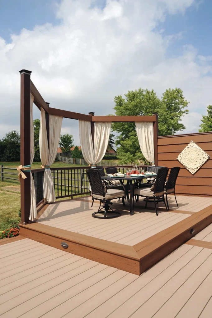 Elevated backyard deck designed with TimberTech Terrain Sandy Birch boards, framed by brown accents and a pergola with flowing cream curtains; the warm, neutral decking color enhances the cozy outdoor dining area and lush green backdrop.