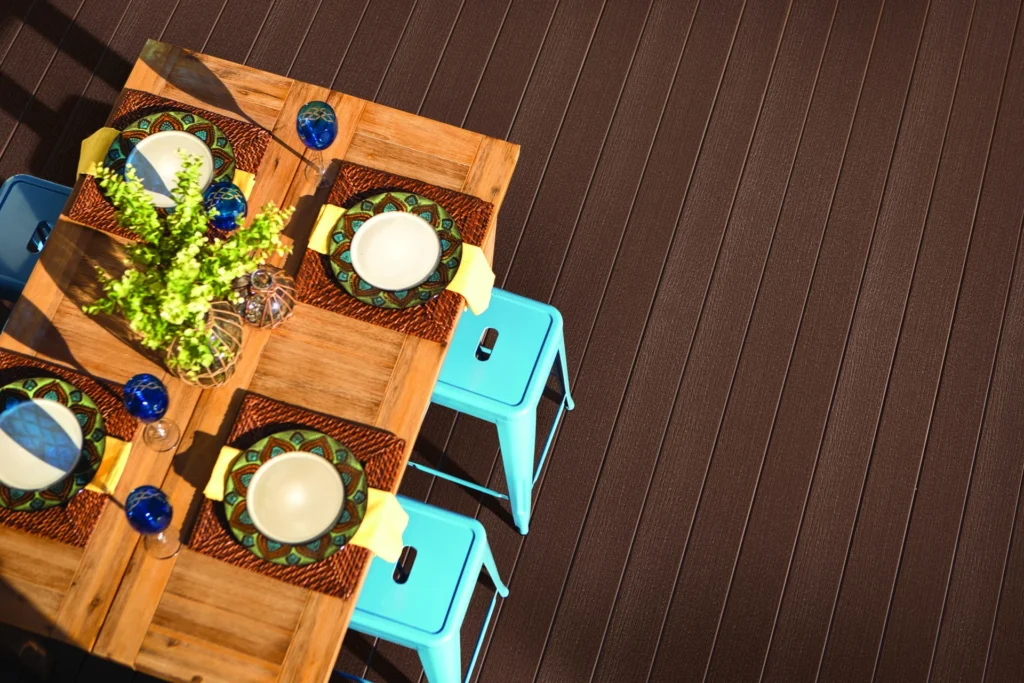 An outdoor dining setup featuring TimberTech Prime composite decking in Dark Teak, shown beneath a rustic wood table and turquoise stools. The decking highlights a deep brown tone with straight wood grain texture, offering a natural look backed by TimberTech’s industry-leading warranties for fade and stain resistance.