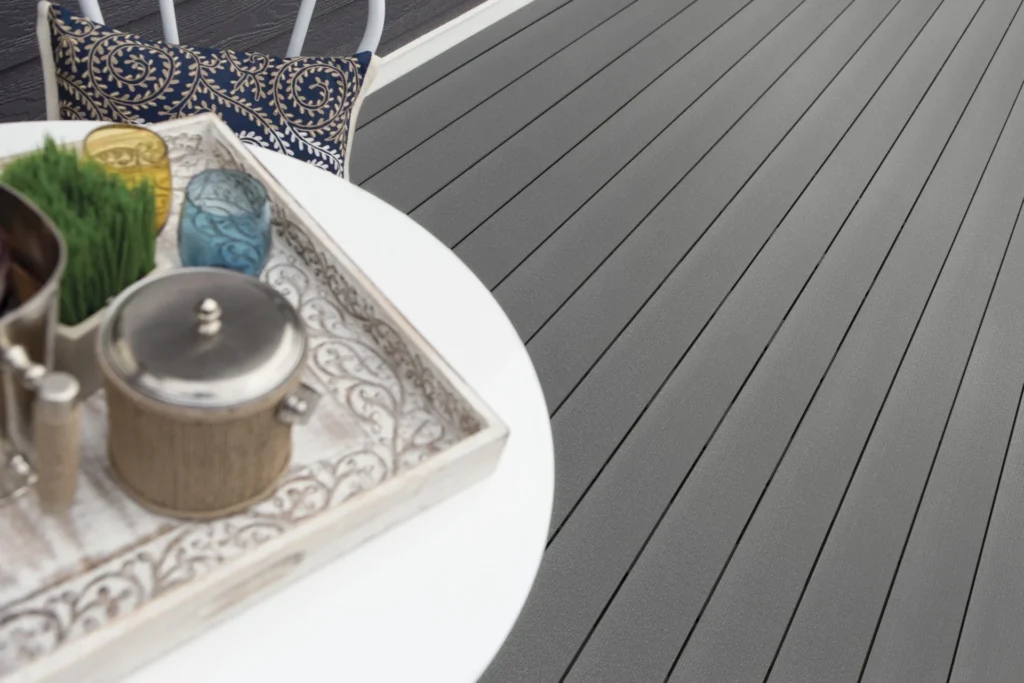 Close-up of TimberTech Prime Maritime Gray composite decking installed on a modern patio. The medium gray boards have a soft, brushed texture and clean lines, creating a contemporary look. A white outdoor table with a decorative tray and colorful glasses sits on the deck, along with a navy cushion with a golden pattern. The subtle grain detailing adds depth to the cool-toned surface, ideal for sleek and stylish outdoor living.