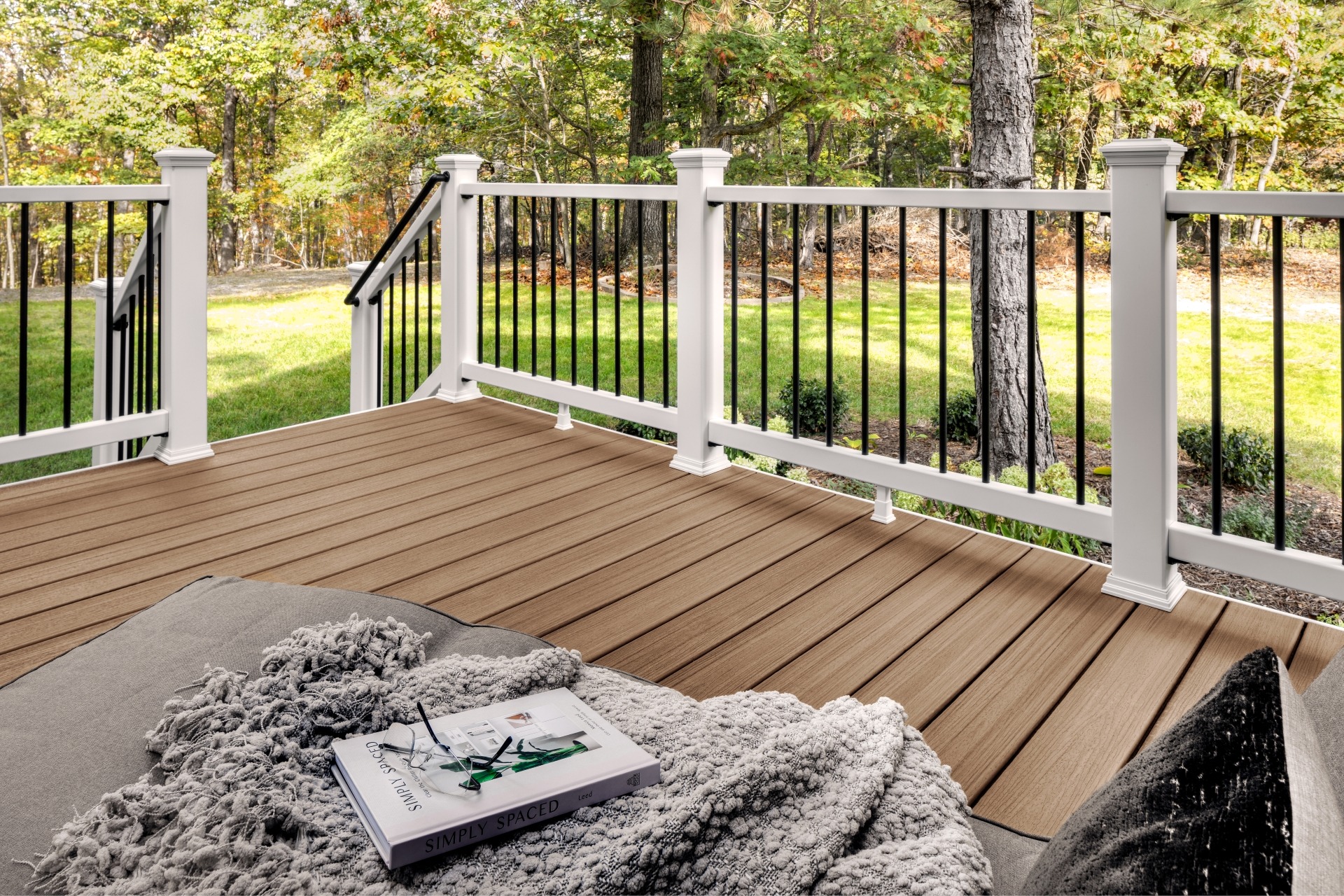 Trex Enhance Golden Hour composite decking with white posts and black balusters overlooking a green backyard, styled with a cozy blanket and book for a relaxed outdoor setting.