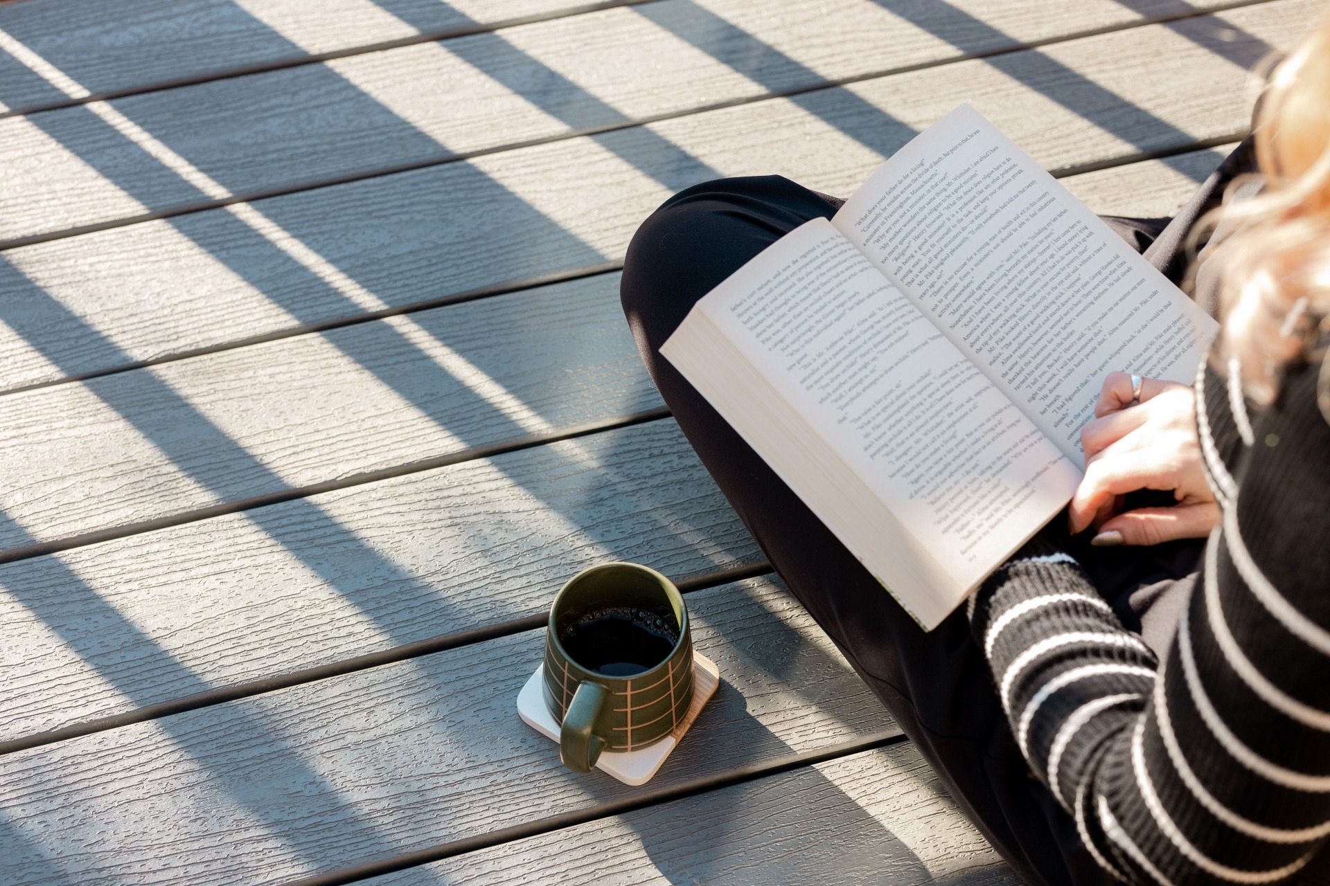 Close-up of Trex Enhance Pebble Beach composite decking with soft gray wood-grain texture, featuring a person reading a book and a cup of coffee resting on the deck in natural sunlight.