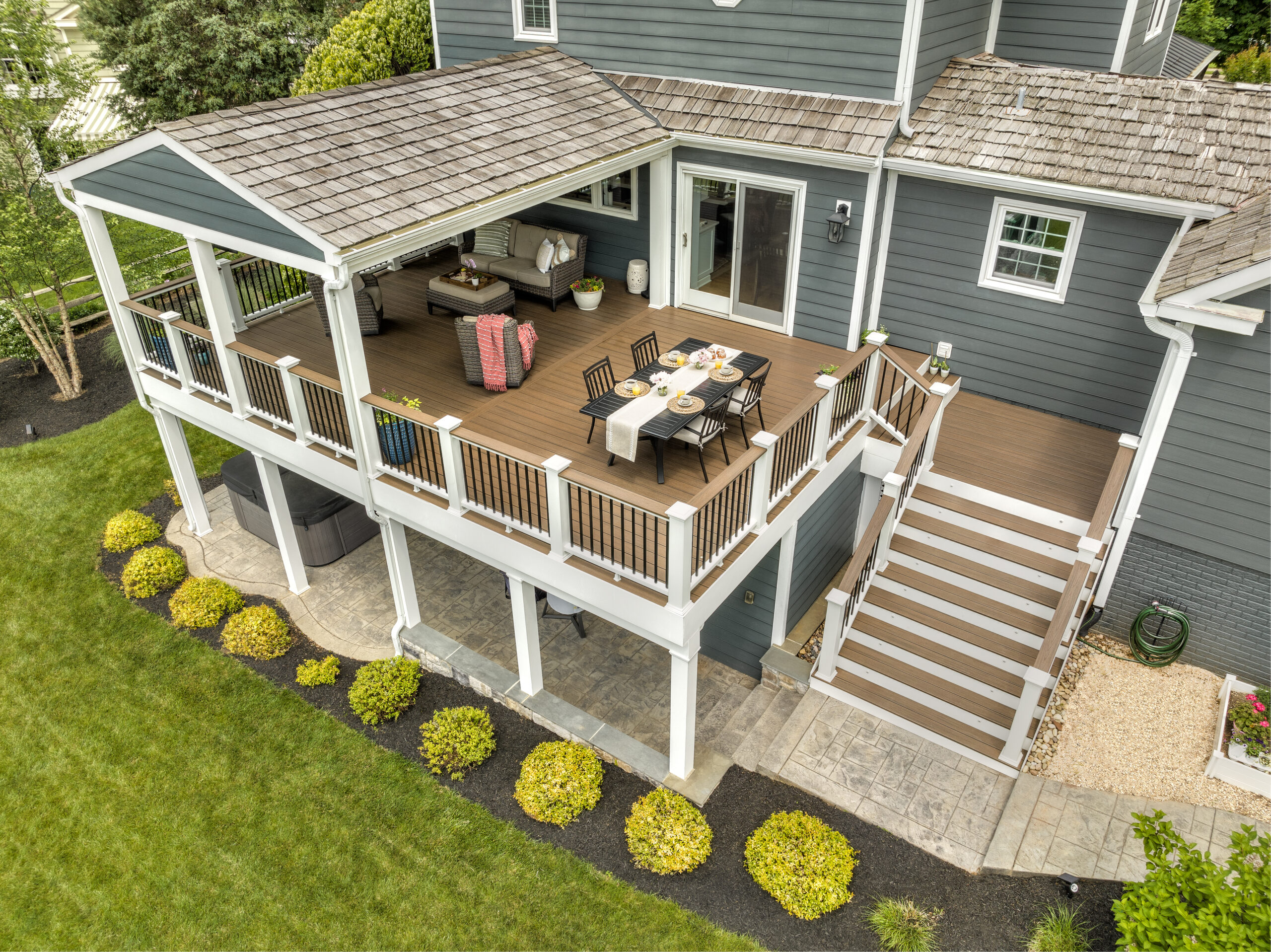 A spacious two-level outdoor deck built with brown Trex composite decking and white railings featuring black balusters. The upper level includes a covered lounge area with seating and a dining table, while wide stairs lead down to a stone patio below. The image highlights consistent deck dimensions, uniform board spacing, and durable composite materials ideal for large outdoor living spaces.