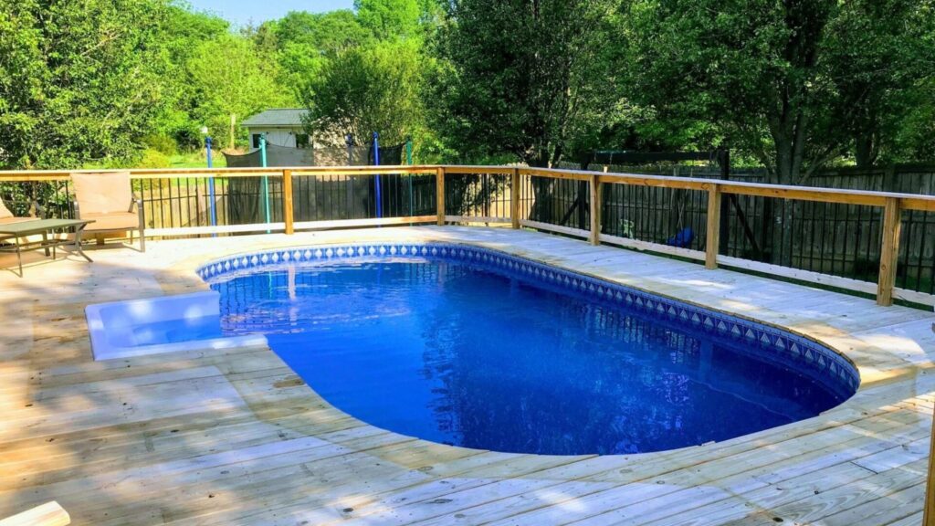 Backyard pool deck with natural wood finish, black railings, and trees in the background.