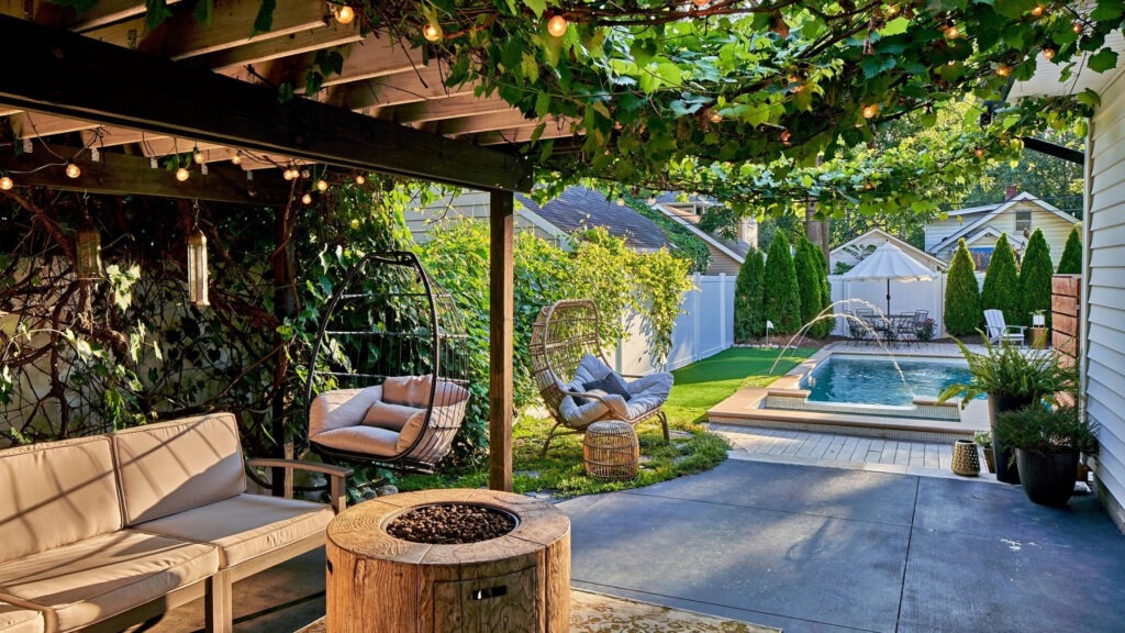 Pergola covered with vines and string lights near pool with cozy chairs and green landscape.