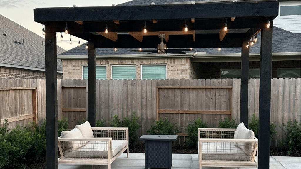 Black wooden pergola with hanging string lights, white cushioned chairs, and privacy fence.