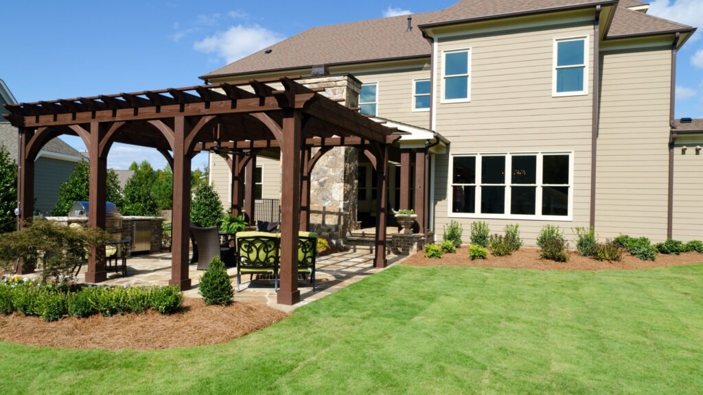 Brown wooden pergola with outdoor kitchen, stone patio, and garden lounge area.
