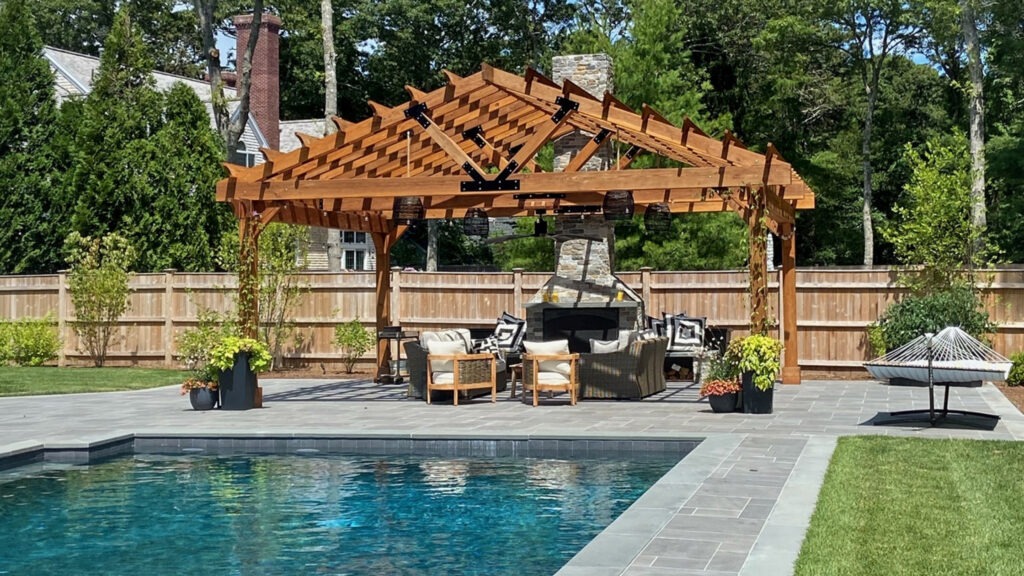 Cedar arched pergola with outdoor fireplace and poolside seating area in backyard.