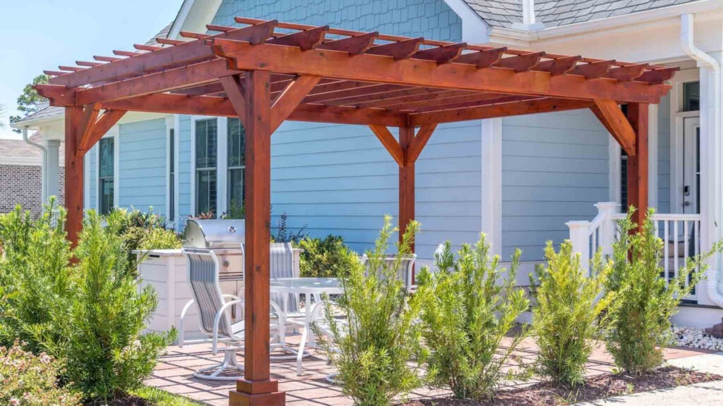 Cedar pergola over patio with outdoor grill, table, and chairs near blue house.