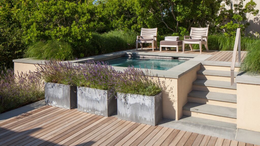 Compact wooden pool deck surrounded by greenery with seating area and small pool.