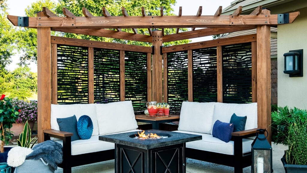 Corner wooden pergola with privacy screens, cushioned sofas, and firepit table surrounded by plants.