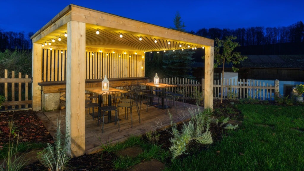 Wooden pergola with string lights, outdoor tables, and garden dining setup at night.