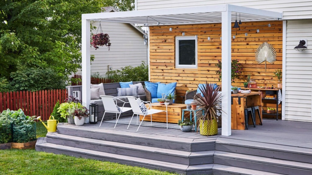 White backyard pergola with wood accent wall gray decking and colorful outdoor seating.
