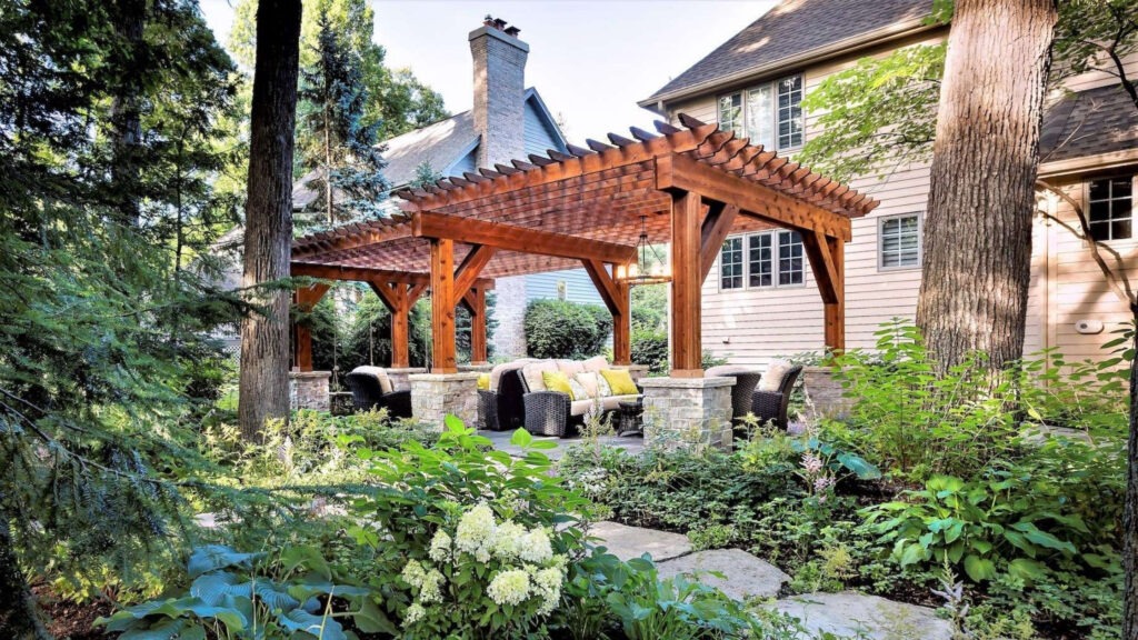Two cedar pergolas with stone columns and wicker seating area surrounded by greenery.