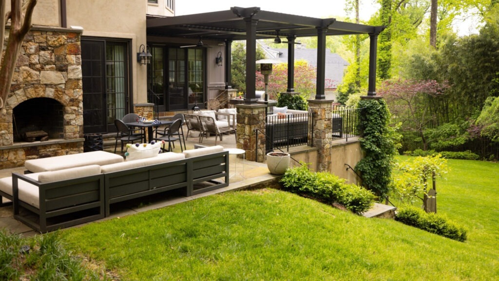 Elevated patio with black pergola, stone columns, outdoor seating, and fireplace overlooking green backyard.