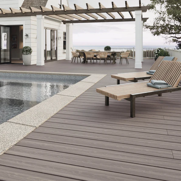A poolside deck built with Fiberon ArmorGuard Coastal Gray, showing medium gray boards with detailed woodgrain and a smooth matte finish. Lounge chairs sit beside the pool, while a pergola and outdoor dining area appear in the background. The color creates a cool, modern, coastal atmosphere.