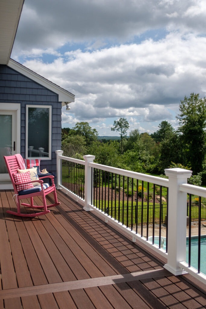 A beautiful second-story deck built with Fiberon Concordia Symmetry Burnt Umber boards, featuring white railings and a cozy pink rocking chair. The deep brown tones of the decking create a warm, inviting atmosphere that complements the blue siding and scenic backyard view.