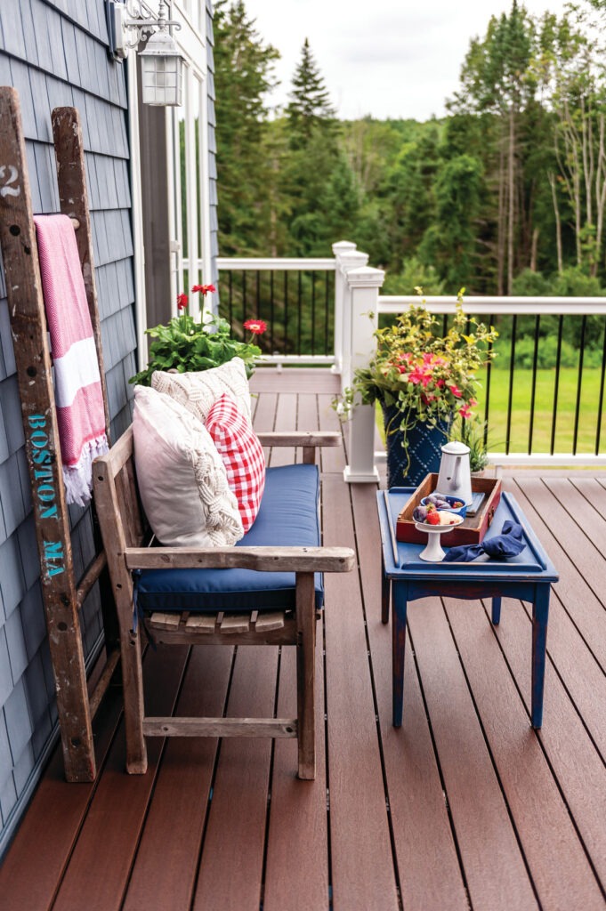 A charming porch setup featuring Fiberon Concordia Symmetry Burnt Umber decking with red and blue accents. The warm brown boards provide a rich, natural contrast against white railings and lush green surroundings.