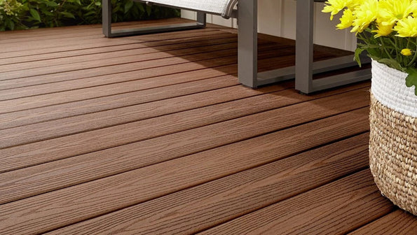 A ground-level view of Fiberon Good Life Bungalow decking installed in a cozy outdoor seating area, displaying its rich brown tones and lightly embossed woodgrain texture. The warm, natural color pairs beautifully with surrounding greenery and modern furnishings. The image emphasizes the board’s durable composite design and long-lasting, low-maintenance finish.