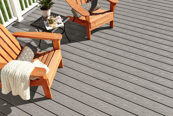 A close-up deck view in the Cottage color, showcasing its even gray hue and fine woodgrain texture under natural sunlight. Wooden lounge chairs and a cozy throw blanket sit on the deck, highlighting the calm, modern look of the solid Weekender Collection finish. The durable capped composite surface appears smooth, stable, and built for long-term performance.
