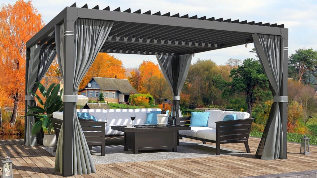 Gray aluminum pergola with curtains, outdoor sectional seating, and fall foliage background.