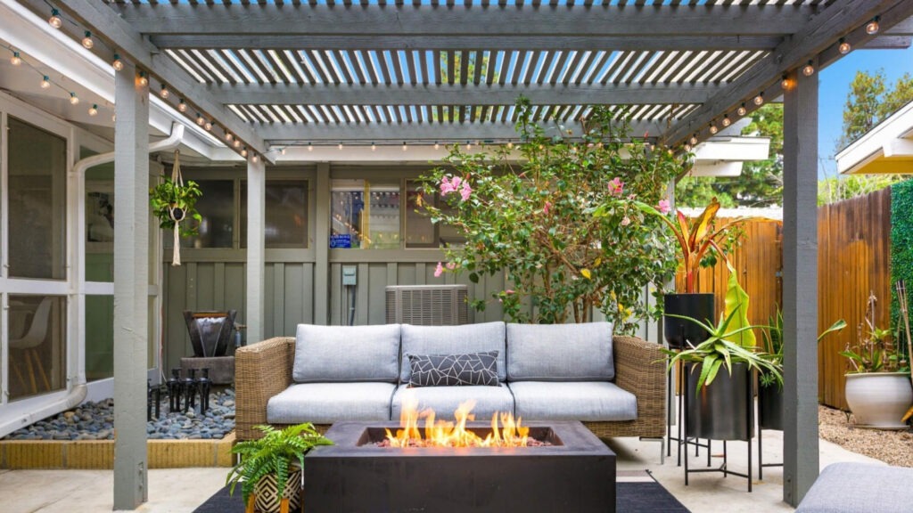 Gray backyard pergola with modern firepit, gray sofa, and potted plants on patio.