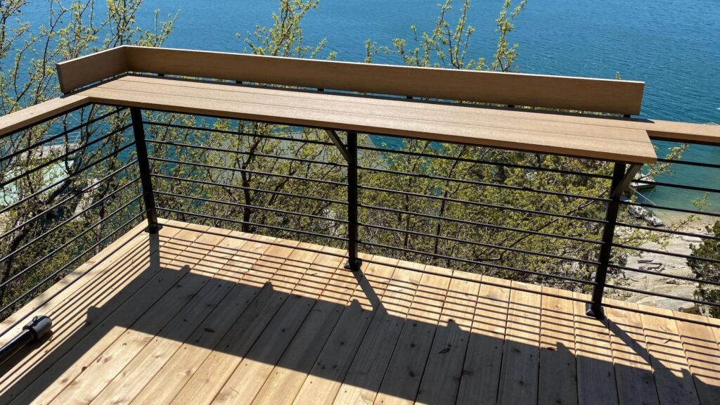 Lakefront wooden deck with black horizontal cable railing and wood top rail overlooking blue water.