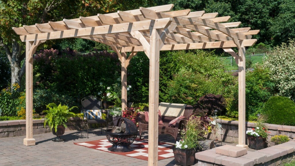 Light cedar pergola with wicker seating, red patterned rug, and firepit in lush garden patio.