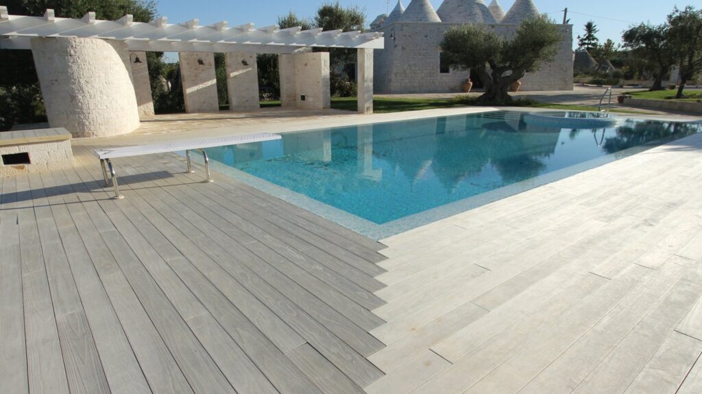 Inground pool with light gray deck, white pergola, and stone architecture.