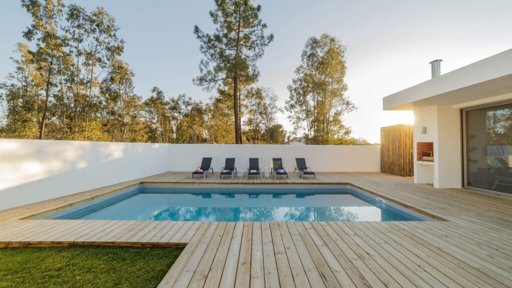 Light wood deck surrounding modern rectangular pool with lounge chairs and white wall.