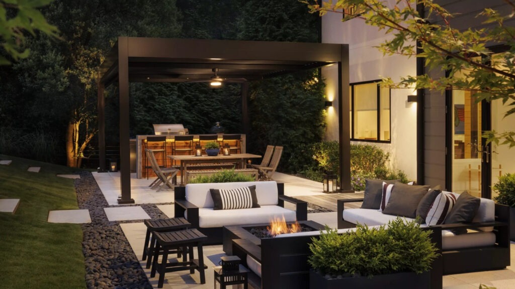 Black metal pergola with outdoor sofa, firepit, dining table, and greenery at night.