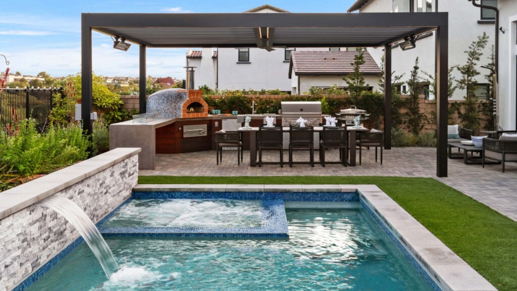 Poolside pergola with outdoor kitchen, pizza oven, bar seating, and modern backyard design.