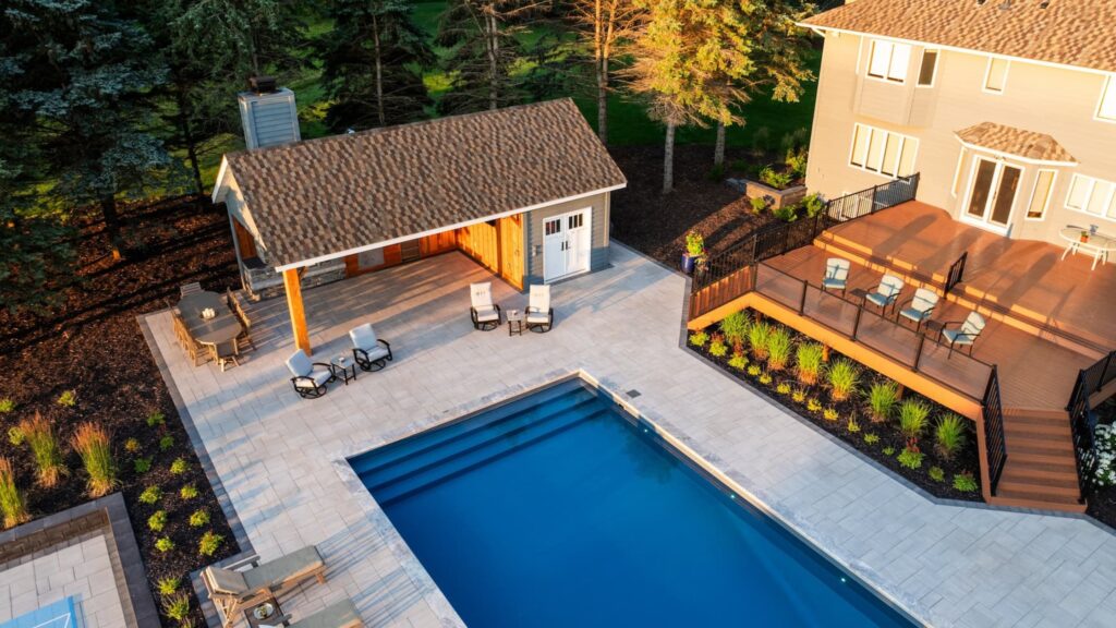 Modern inground pool with raised wood deck, outdoor kitchen, and patio seating area.