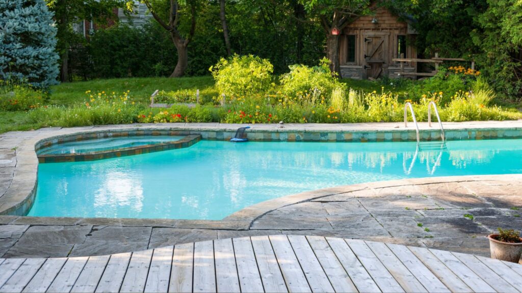 Inground pool with curved stone patio and wood deck surrounded by lush garden landscaping.