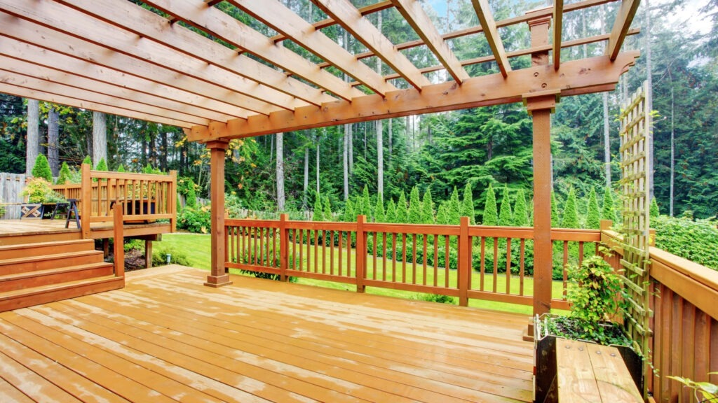 Wooden pergola on raised backyard deck with railing and garden trees in the background.