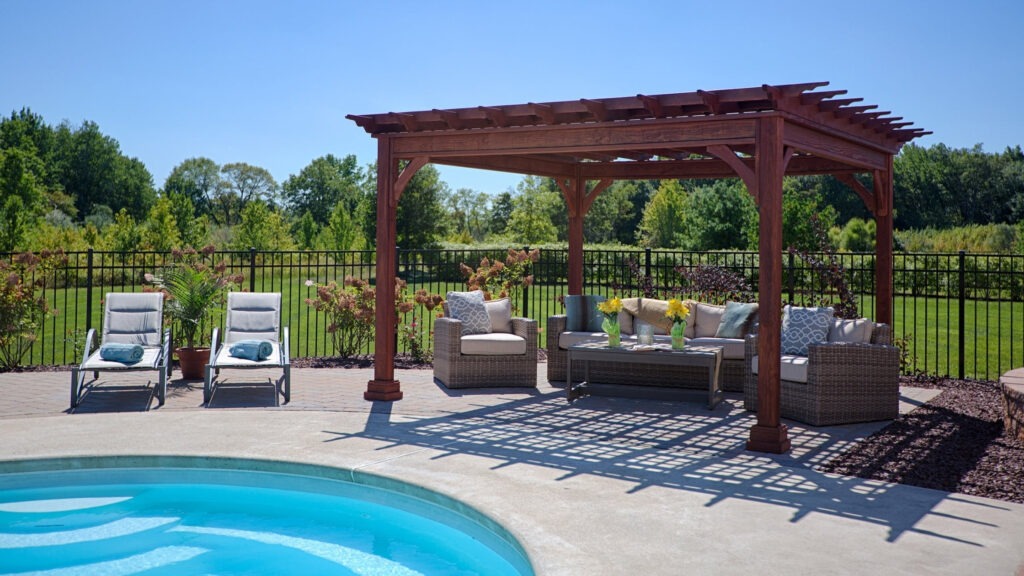 Cedar pergola by pool with wicker patio furniture and open green backyard view.