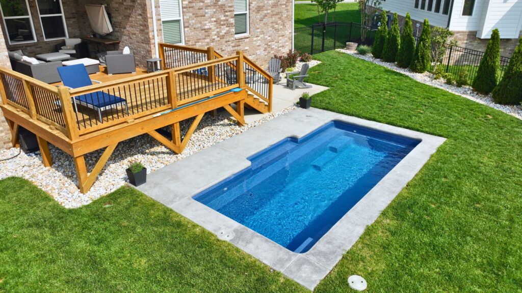 Elevated wood deck with lounge seating above a rectangular inground pool surrounded by grass.